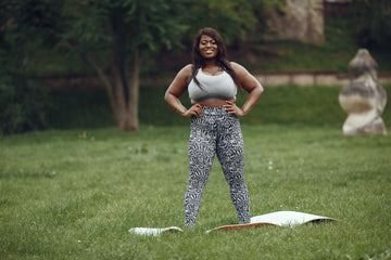 Confident plus-size woman in activewear leggings posing outdoors on grass with yoga mat