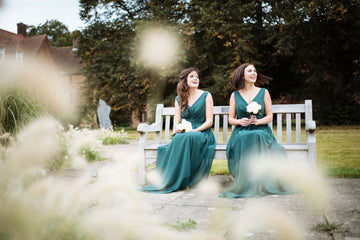 Two plus size bridesmaids in emerald green dresses smiling together on a garden bench